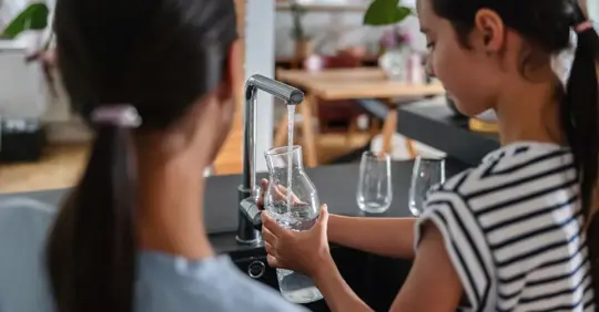 Young Sisters Filling A Glass Jar With Some Tap Water At The Kitchen